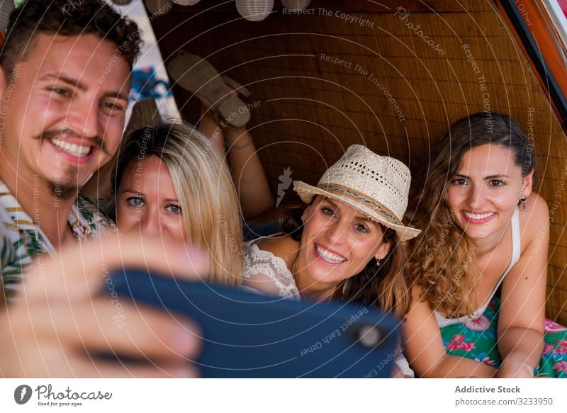 Smiling man taking selfie with girlfriends comfortable placing on trunk of blue car people smartphone seaside using music travel minivan having fun gadget