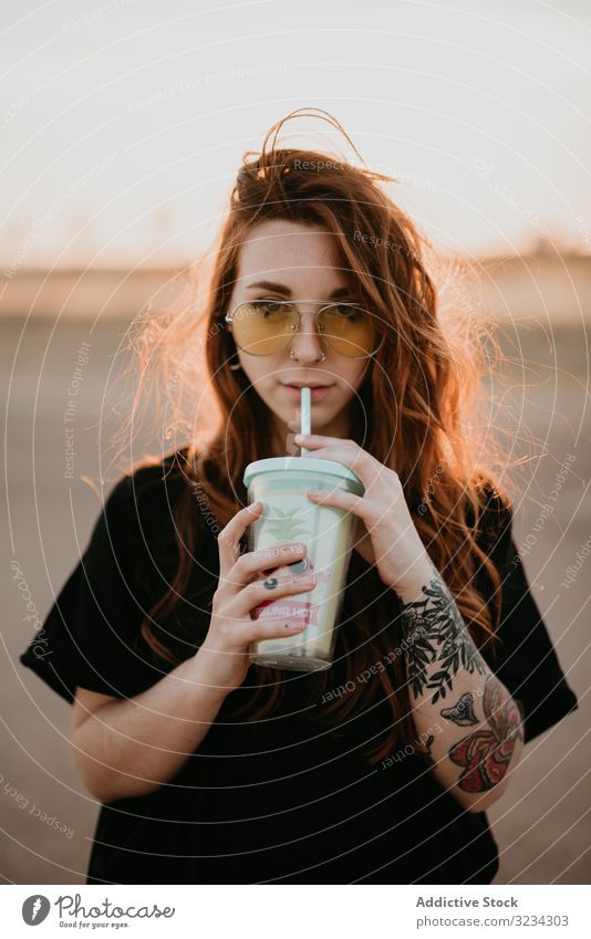 Long haired teenage girl drinking milkshake and looking at camera woman millennial cheerful cool style happy sunset tattoo sunglasses piercing leisure smile
