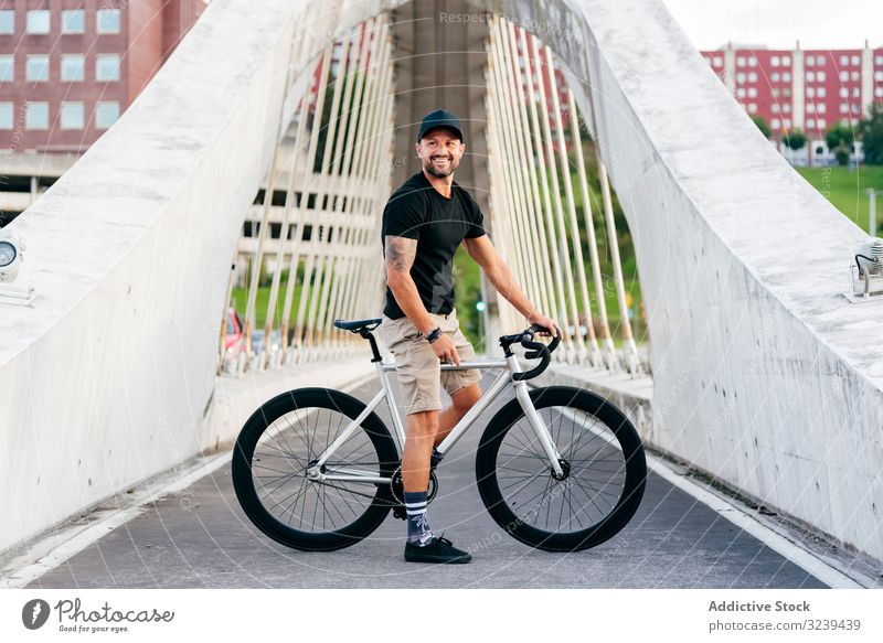 Cheerful man with bicycle standing on footbridge bike city ride modern active sportive summer male adult happy cheerful smile beard cap cyclist recreation