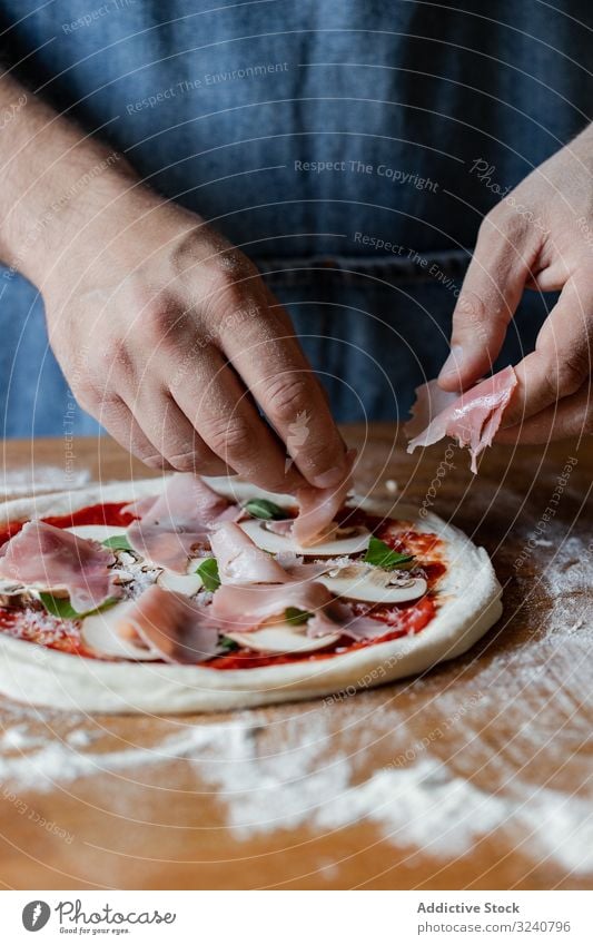 Crop man putting meat on pizza ham dough flour soft fresh cuisine kitchen preparation food cheese tomato mushroom herb sauce chef italian male apron raw