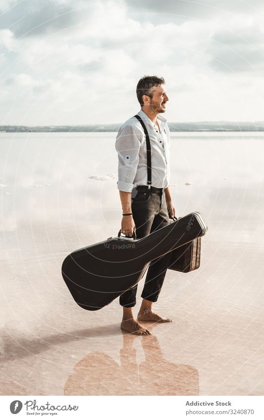 Focused stylish man with suitcase and guitar gig bag on seaside serious barefoot carry water usa shore gazing contemplation modern artist musician young adult