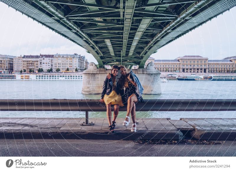 Smiling girlfriends hugging and sitting on fence along river under bridge women travel city tourist architecture fun smiling vacation together tourism budapest