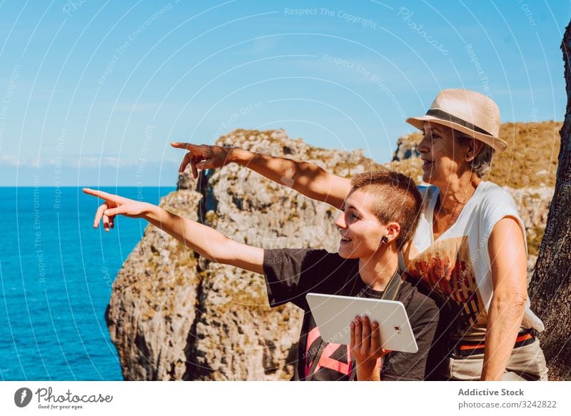 Stylish woman with tablet in hands and elderly lady pointing with finger to blue sea women mature rocky using water sky vacation summer ocean happy tropical
