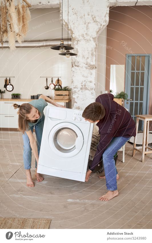 Enthusiastic couple carrying washing machine into new kitchen moving furnish apartment appliance home domestic cheerful delivery property together partnership