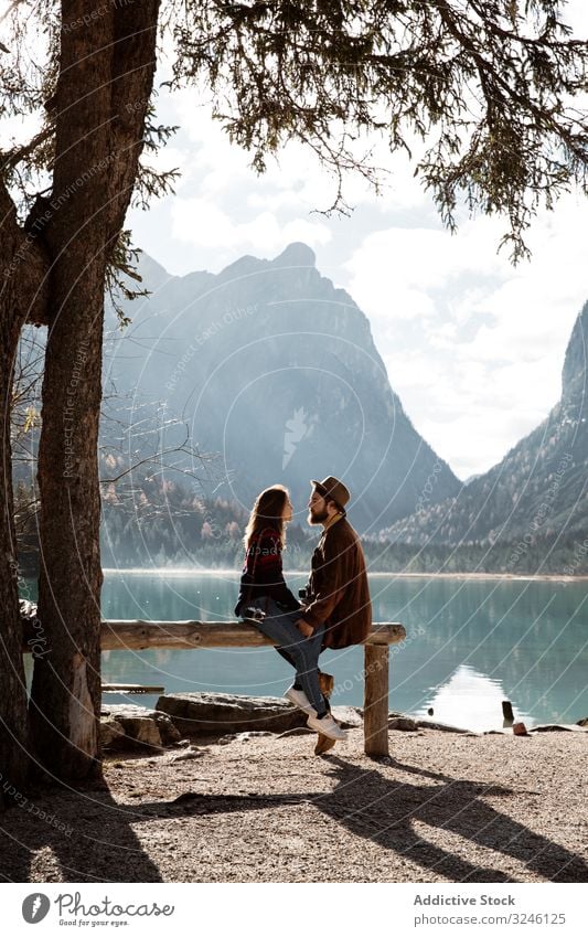 Male and female travelers sitting on fence near lake and mountains couple nature shore pine tree big sunlight holding hands wooden summer adventure lifestyle