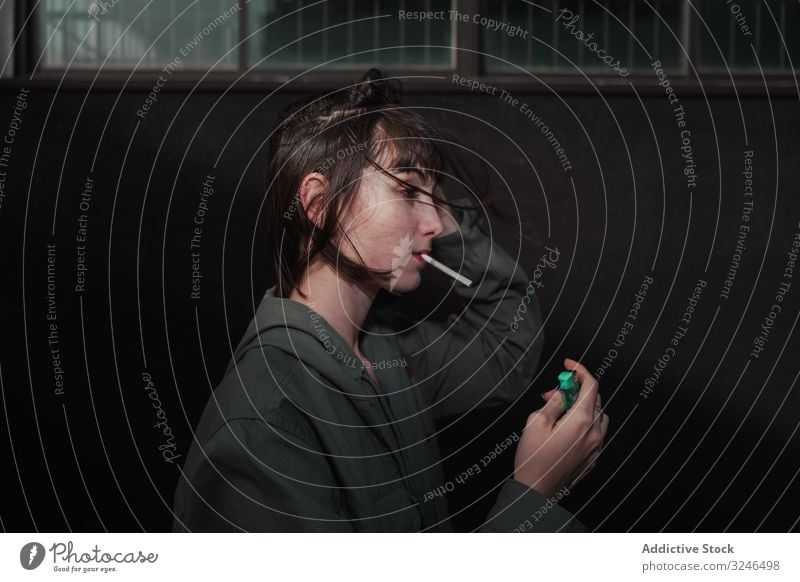 Female teenager in casual clothing smoking cigarette at city street light up holding liter wear jacket outfit grey wall nail blue brunette rebel sad modern
