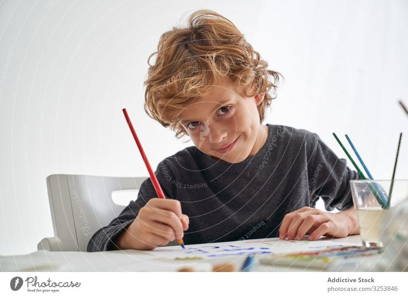 Cheerful boy painting and looking at camera smile paintbrush home art creative picture education table sit kid child tool craft study learn elementary