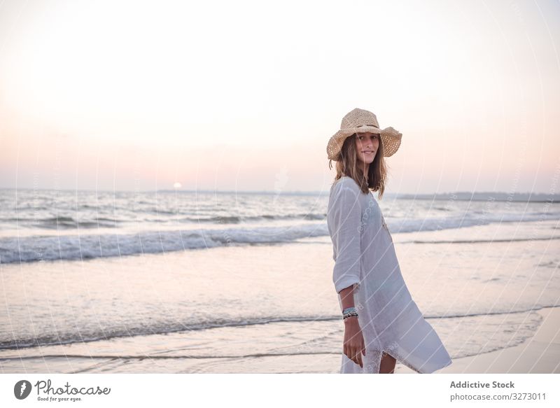 Charming woman in light white dress looking at camera over shoulder on wavy beach sea traveling leaning seaside tourism charming vacation hat curly holiday