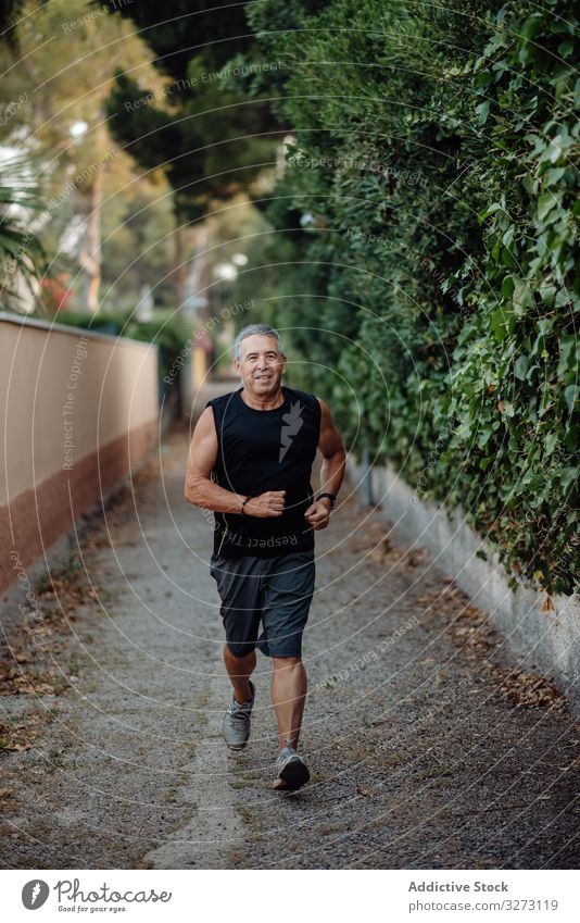Old man in good shape running on road along plant fence sportive jogging elderly street mature old training runner athlete workout fitness city healthy athletic