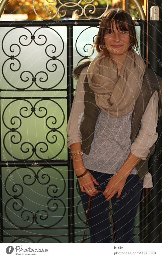 Young fashionable, happy, smiling young woman; teenager with scarf and pony, standing in the shade in front of a wrought-iron gate of a garden and taking a break during the walk.