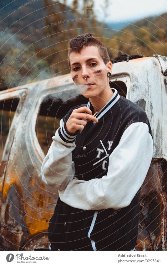 Relaxed woman with cigarette leaning on burnt car smoke rebel rest rusty countryside young lifestyle freedom vehicle auto androgynous short hair subculture