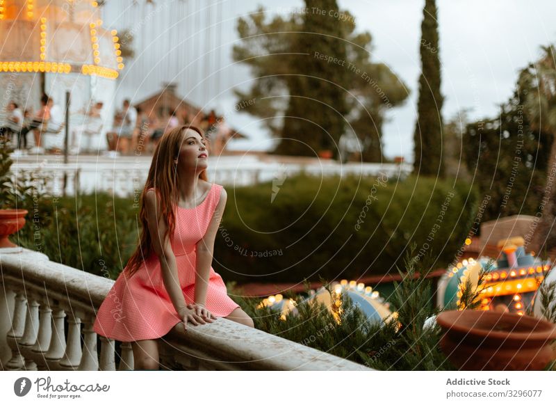 Young woman in pink dress sitting on fence in amusement park funfair romantic sensual carousel teenager fashion entertainment style trendy millennial relax