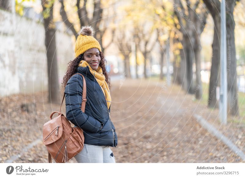 Stylish content black woman in hat and jacket with modern backpack looking at camera in park leaves alley autumn nature stylish african american natural street