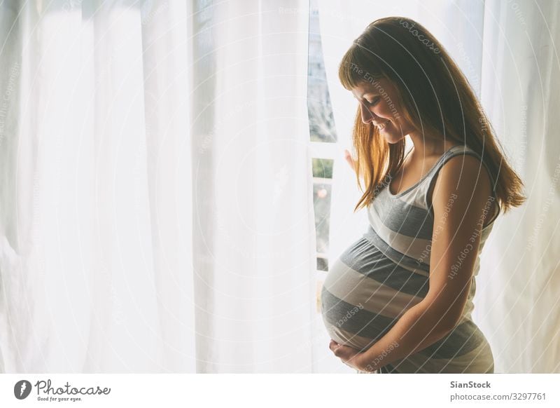 Young beautiful pregnant woman standing near window at home - a