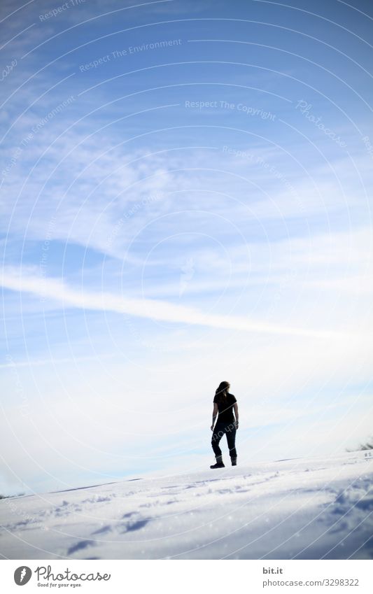 Sporty young woman in black clothing stands sleeveless, on a mountain in the snow in winter and looks up at the blue sky. Rear view of a female hiker, mountaineer, who has left tracks in the snow to enjoy the view down to the valley.