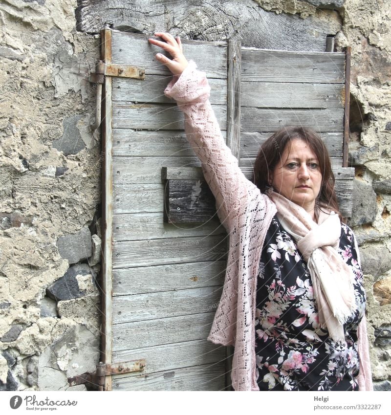 long-haired brunette woman with patterned dress, pink jacket and pink scarf is standing in front of a wall with old closed wooden window Human being Feminine