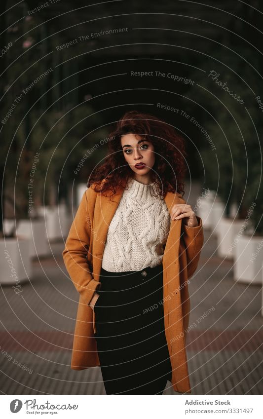 Young woman in overcoat with arms raised looking at camera with provocative eyes young standing curly haired challenge jumper knitted building warm female