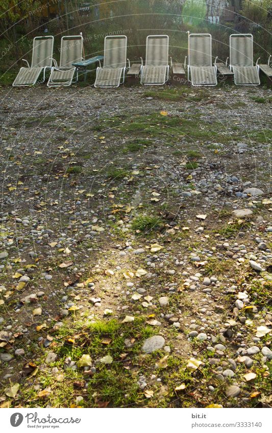 Old, broken, unkempt, white, empty plastic deck chairs, standing next to each other in a row, in the light on a mooring with grass and stones, waiting for holidaymakers / visitors.