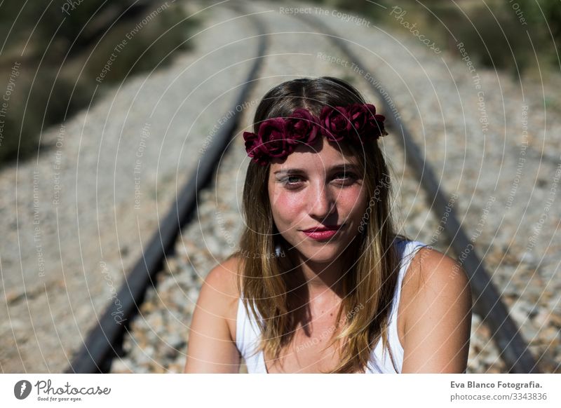 portrait of a young beautiful woman wearing a red roses wreath on her head and looking at the camera.Sitting on a railway. Outdoors. Sunny. Lifestyle Freedom