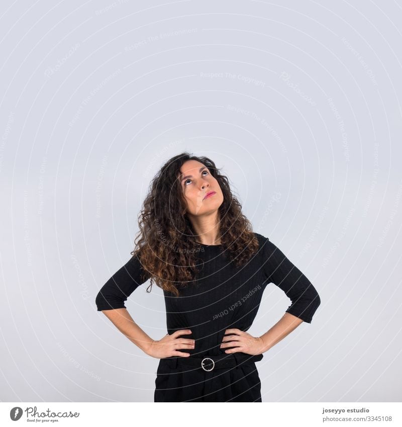 Curly haired brunette woman dressed in black with her hands on her waist looking up with thoughtful gesture. 30-40 years advertisement attitude background