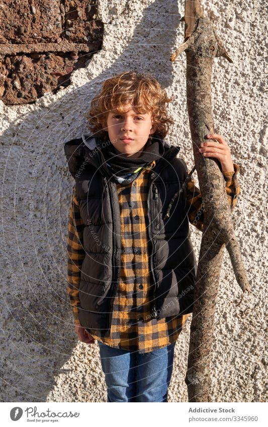 Adorable boy in warm vest holding tree trunk and looking at camera rural explorer warm clothes adventurer village child wall tourist holiday kid childhood