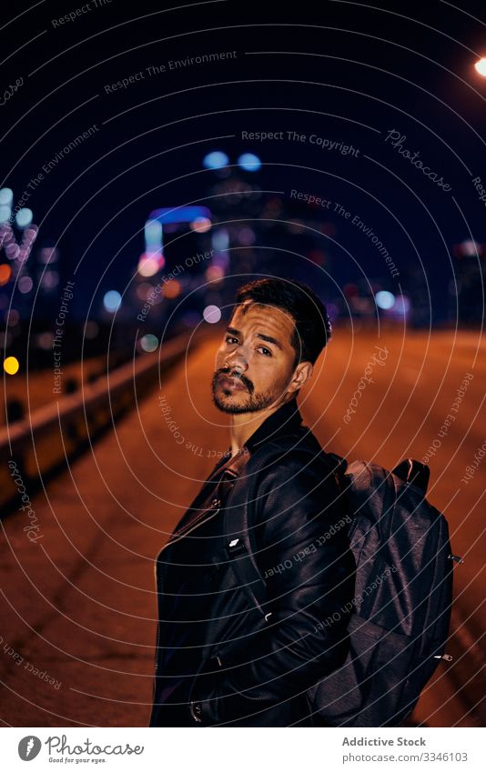 Confident Hispanic guy in stylish clothing at night city man street sidewalk backpack jacket leather urban modern male lifestyle young downtown handsome dusk