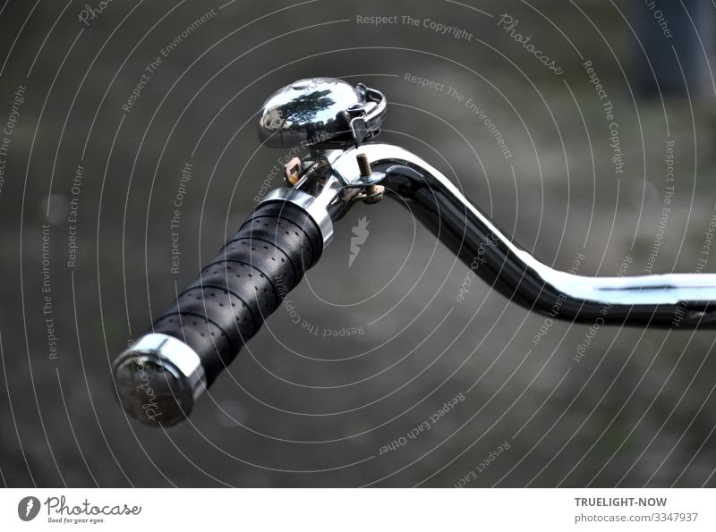 In front of a blurred background a part of a chromed retro bicycle handlebar with a perforated leather grip and a shiny bell, in which the surroundings are reflected, glows