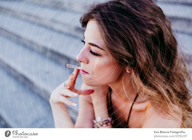 Beautiful young caucasian woman sitting on stairs at the city street on a sunny day and smoking a cigarette. Urban lifestyle and smoking concept. close up view