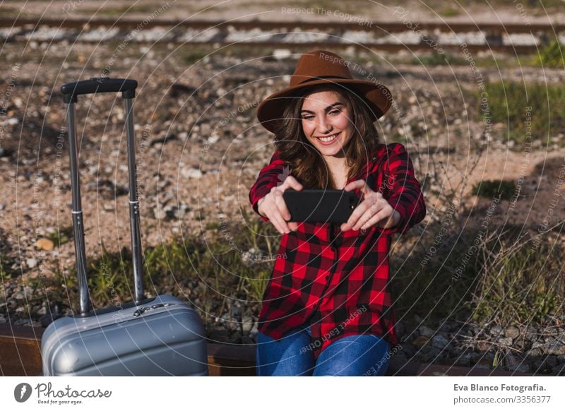 young beautiful woman wearing casual clothes, walking by the railway with suitcase and mobile phone and smiling. Outdoors lifestyle. Travel concept. She is taking a picture or selfie