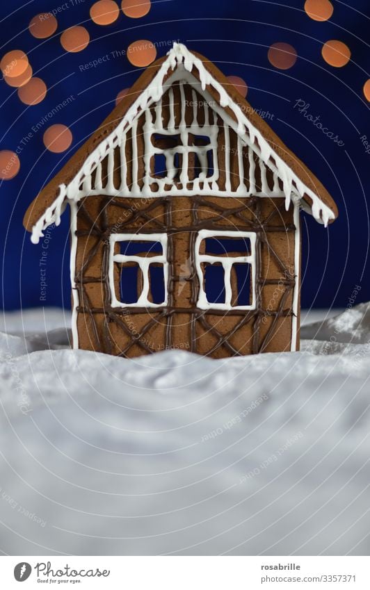 Gingerbread house facade with icing and chocolate decoration as half-timbered on white velvet, which looks like snow, in front of a dark blue background with bokeh dots as stars