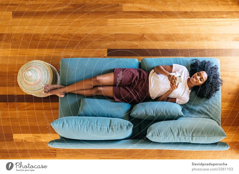 Black woman chatting on smartphone lying on couch at home using chill lounge sofa living room modern relax african american black rest browsing mobile smile