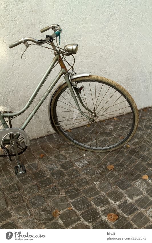 alt l old, nostalgic wheel in retro style, parked in front of a white, bright house wall on cobblestone. Front part, handlebar, lamp, light, tires, brake of grey wheel parked in the city. Risk of theft of old bike, not locked.