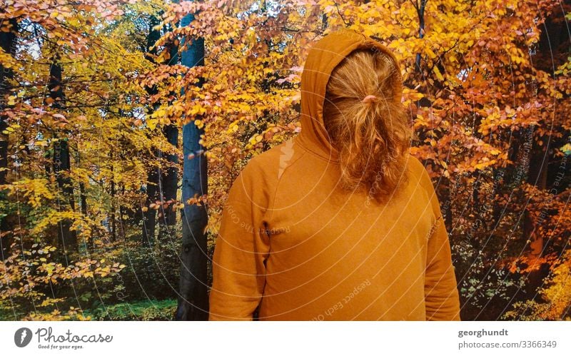 Man has tied his long red hair in front of his face in a braid. He is wearing a brown-orange hooded sweater and is standing in front of a large photo of an autumn forest.
