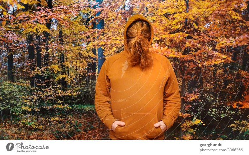 Man has tied his long red hair in front of his face in a braid. He is wearing a brown-orange hooded sweater and is standing in front of a large photo of an autumn forest.