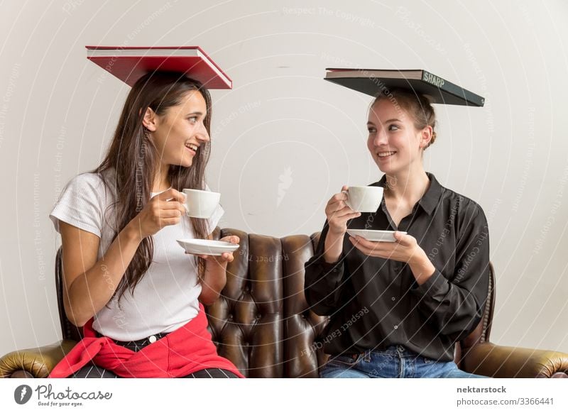 Two Young Women Balancing Books on Head and Holding Coffee Cups Making Eye Contact females eye contact looking at each other girl women young adult