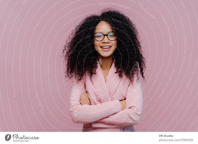 Pleased curly haired woman wears optical glasses for vision correction, elegant suit, keeps hands crossed over chest, poses against purple background, comes on job interview. Businesswoman at work