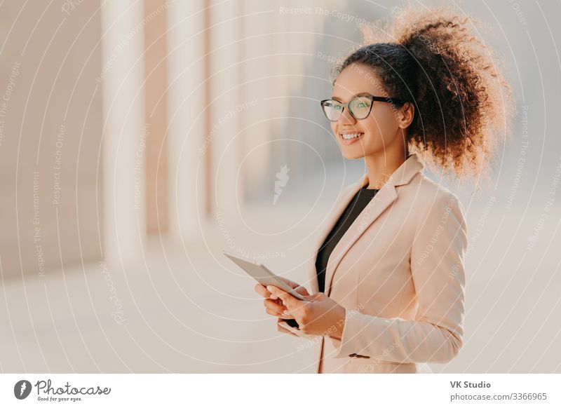 Profile shot of glad dark skinned woman with Afro hair, dressed elegantly holds tablet computer in hands strolls at street wears optical glasses connected to high speed internet. Lifestyle, technology