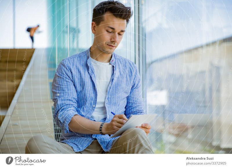 Man writing in notebook, sitting on steps at big windows, big working space. adult casual checklist concentrated concept creative creativity designer diary