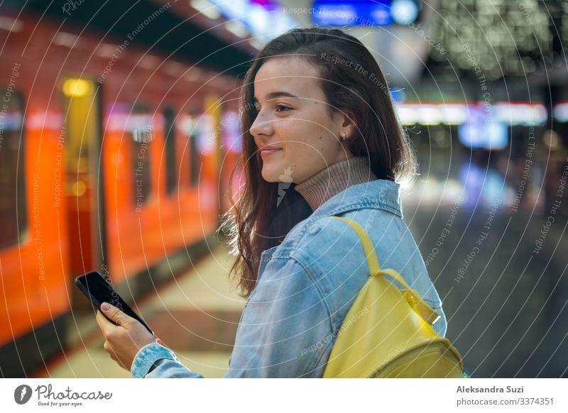 Teenager girl in jeans with backpack standing on metro station holding smart phone in hand, scrolling and texting, smiling and laughing. Futuristic bright subway station. Finland, Espoo