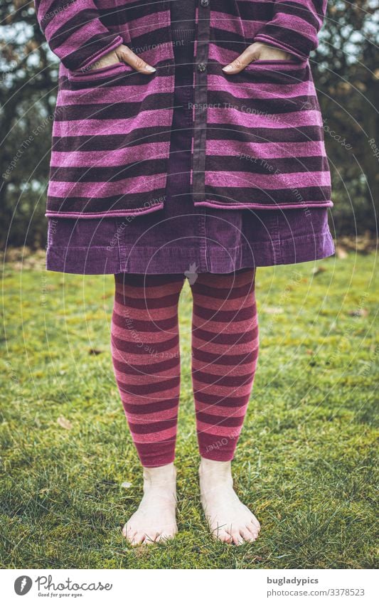 A female person stands barefoot on grass, in the background there is a hedge. The peron can be seen from chest height to the feet. She is wearing a purple striped cardigan, a purple cord skirt and pink striped leggings.