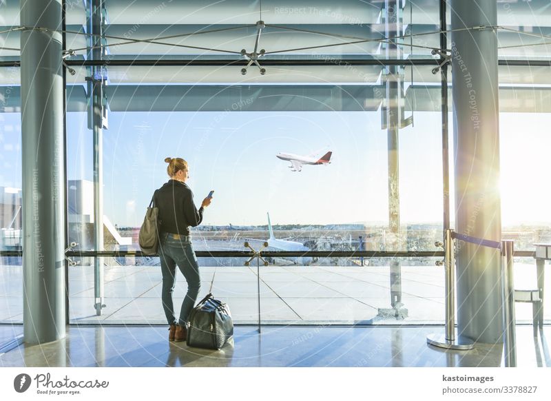 Young casual female traveler at airport, holding smart phone device, looking through the airport gate windows at planes on airport runway. Vacation & Travel