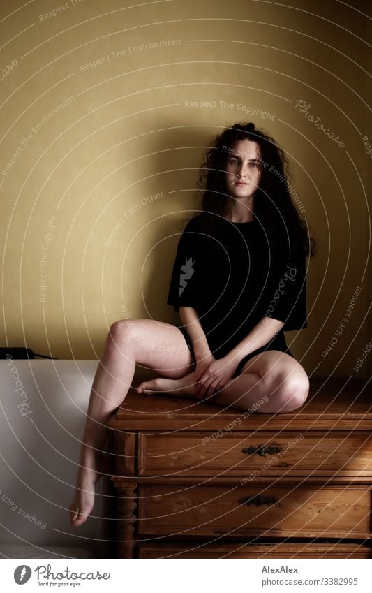 Young woman sitting on a chest of drawers in front of a yellow