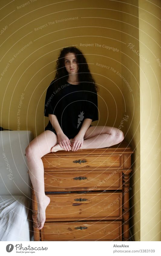 Young woman sitting on a chest of drawers in front of a yellow wall Looking into the camera Portrait photograph Central perspective Shallow depth of field Day