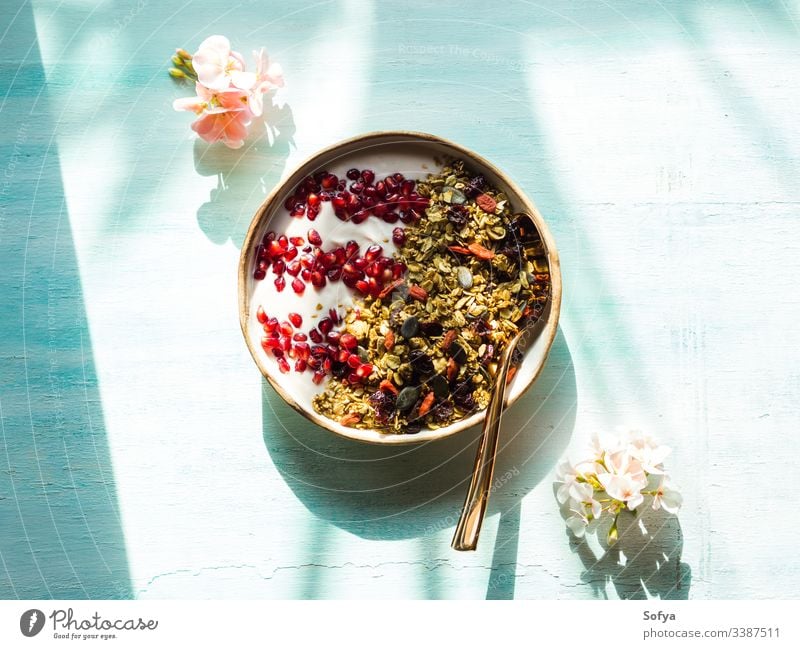 Home made granola bowl with pomegranate seeds, matcha granola with oats and dried fruit, nuts on turquoise background with pink flowers. breakfast delicious