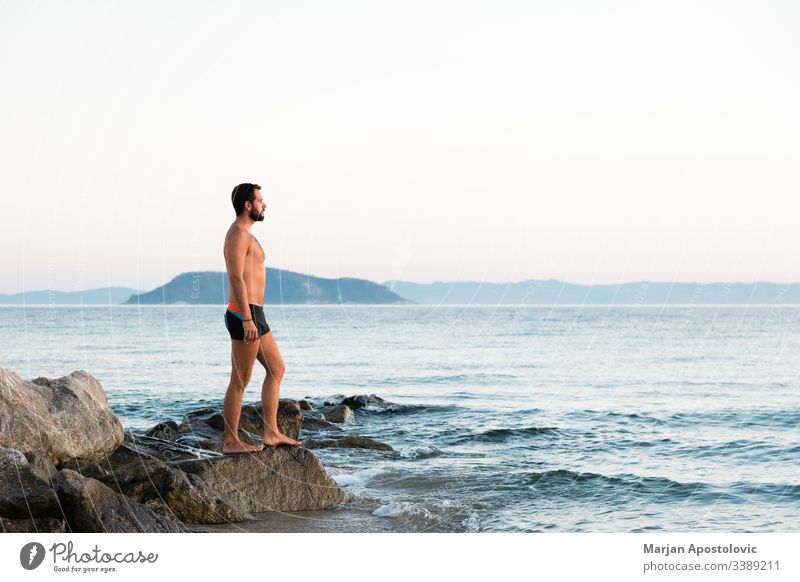 Young man standing on the rock enjoying the sunset by the sea adult alone background beach calm caucasian coast dusk evening free freedom handsome healthy