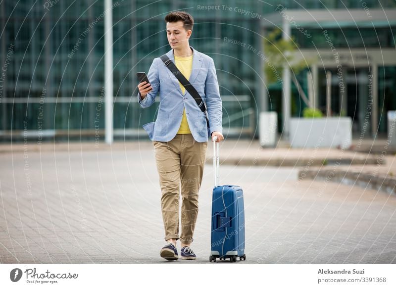 Stylish man standing at airport with smartphone and suitcase, browsing, texting, using mobile app. Business traveling. application arrival bag business