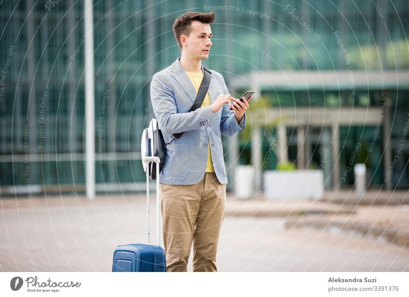 Stylish man standing at airport with smartphone and suitcase, browsing, texting, using mobile app. Business traveling. application arrival bag business