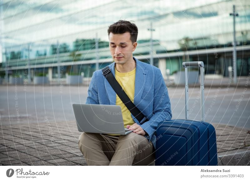 Stylish man sitting at airport with suitcase and laptop, working, typing, browsing. Businessman traveling. adult bag baggage business businessman busy caucasian