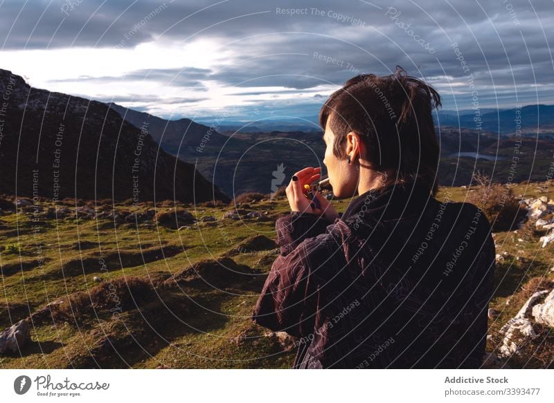 Cool young woman lighting cigarette standing against amazing landscape of mountain ridge in cloudy weather highland smoke rebel travel cool escape hipster fire