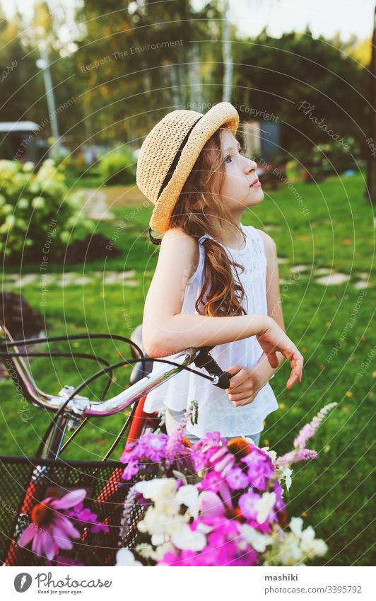 happy child girl in hat riding bicycle with bouquet of wild flowers, summer vacations concept book kid childhood outdoor nature park fun lifestyle read leisure